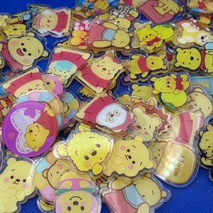 May include: A collection of colorful Winnie the Pooh character acrylic charms. The charms feature various poses and designs of the beloved bear and his friends, with yellow, red, and pink accents. The charms are clear and shiny.