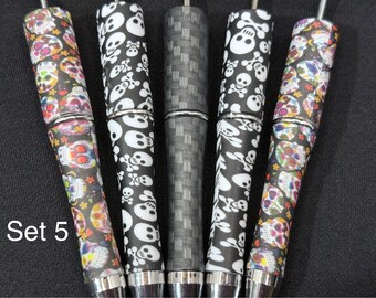 May include: Set of five pens with decorative barrels. Two pens feature colorful sugar skull designs, two have black and white skull and crossbones patterns, and one has a gray woven pattern. The pens have silver-colored metal accents.