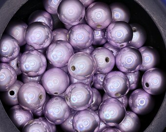 May include: A collection of iridescent lavender-colored beads. The round beads have a glossy finish and a hole drilled through the center. The beads are piled together in a dark container, creating a reflective surface.