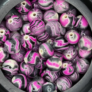 May include: A collection of round beads with a swirled pattern in shades of pink, black, and white. The beads are contained within a dark container. These beads are suitable for jewelry making and crafting projects.