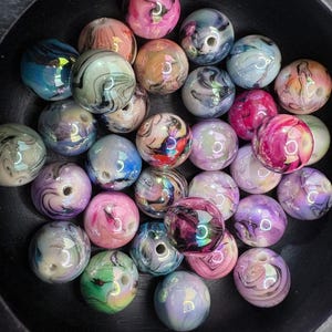 May include: A collection of colorful, marbled beads in a variety of colors including pink, blue, purple, and green. The beads have a glossy finish and are arranged in a black bowl. Each bead has a hole for stringing.