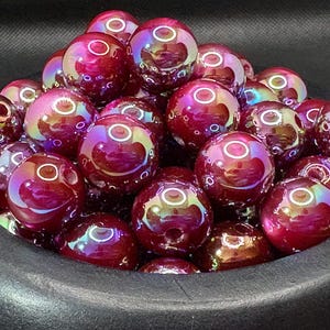 May include: A collection of iridescent, deep red glass beads with a glossy finish. Each bead has a hole for stringing and reflects a rainbow of colors. The beads are round and appear to be approximately 0.75 inches in diameter.