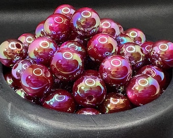 May include: A collection of iridescent, deep red glass beads with a glossy finish. Each bead has a hole for stringing and reflects a rainbow of colors. The beads are round and appear to be approximately 0.75 inches in diameter.