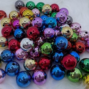 May include: A collection of shiny, metallic beads in various colors, including gold, silver, red, blue, green, and purple. Each bead has a reflective surface and a small hole. The beads are scattered on a white, fluffy surface.
