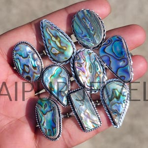 May include: A collection of silver rings with iridescent abalone shell stones in various shapes, including oval, teardrop, and rectangular. The rings are displayed on a hand, showcasing the natural colors of the shells.