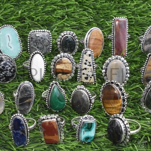 May include: A collection of 20 silver rings with various natural stone inlays. The stones are in a variety of shapes, sizes, and colors, including green, brown, black, white, and blue. The rings are set on a green grassy background.