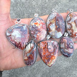 May include: A collection of seven gemstone pendants in various shapes, including oval and teardrop. The pendants feature intricate patterns in shades of red, brown, and cream, set in silver-toned metal. Each pendant has a loop for a chain.