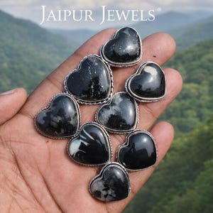 Op de afbeelding: Een hand houdt meerdere hartvormige edelstenen met zilverkleurige randen vast. De stenen zijn voornamelijk zwart met witte adering. De achtergrond toont een bergketen en de tekst "JAIPUR JEWELS".