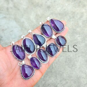 May include: A collection of amethyst gemstone pendants in various shapes, including oval, teardrop, and round. Each pendant has a silver-colored metal setting with a detailed border. The polished purple stones are displayed against a neutral background.