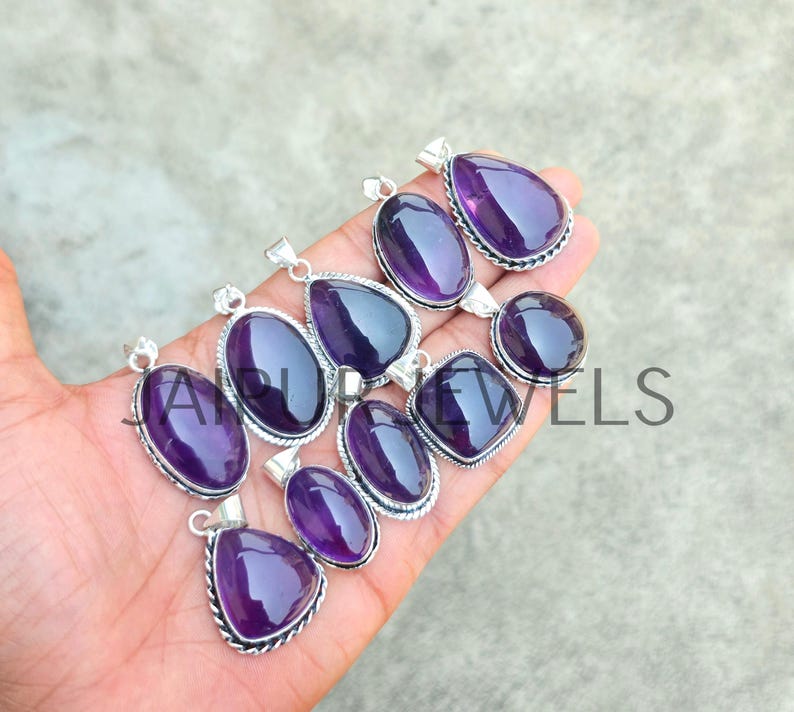 May include: A collection of amethyst gemstone pendants in various shapes, including oval, teardrop, and round. Each pendant has a silver-colored metal setting with a detailed border. The polished purple stones are displayed against a neutral background.