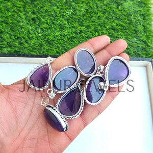 May include: A collection of amethyst pendants in various shapes, including teardrop and oval, set in silver-toned metal. The pendants are displayed on a hand against a backdrop of green grass. The text "JAIPUR JEWELS" is visible.