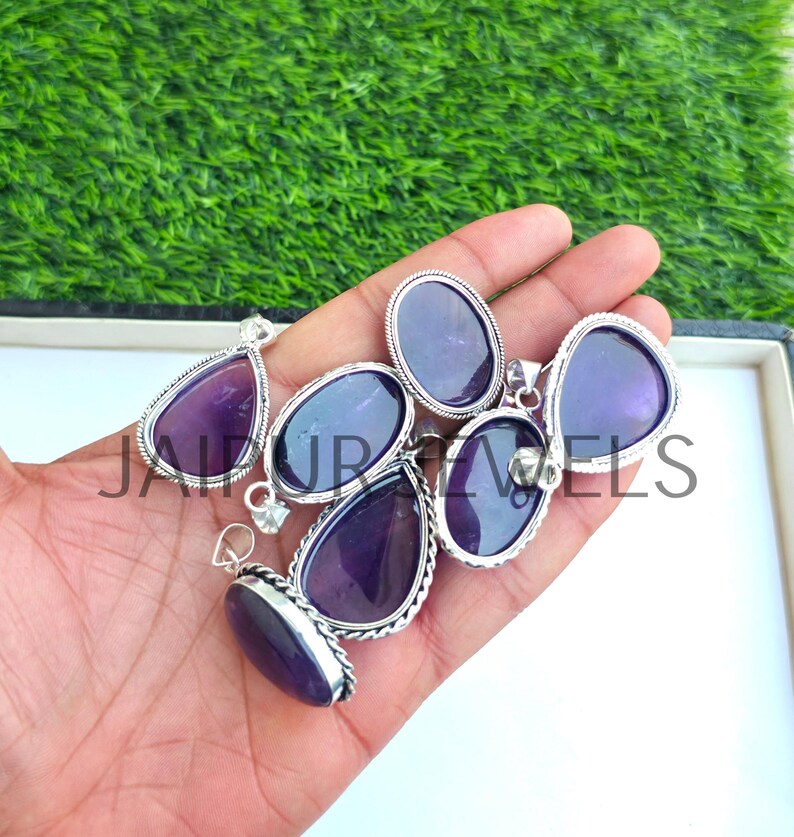 May include: A collection of amethyst pendants in various shapes, including teardrop and oval, set in silver-toned metal. The pendants are displayed on a hand against a backdrop of green grass. The text "JAIPUR JEWELS" is visible.