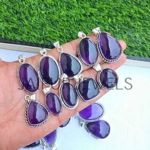 May include: A collection of amethyst pendants in various shapes, including oval, teardrop, and square. Each pendant features a polished, deep purple gemstone set in a silver-colored metal frame. The pendants are displayed against a green background.