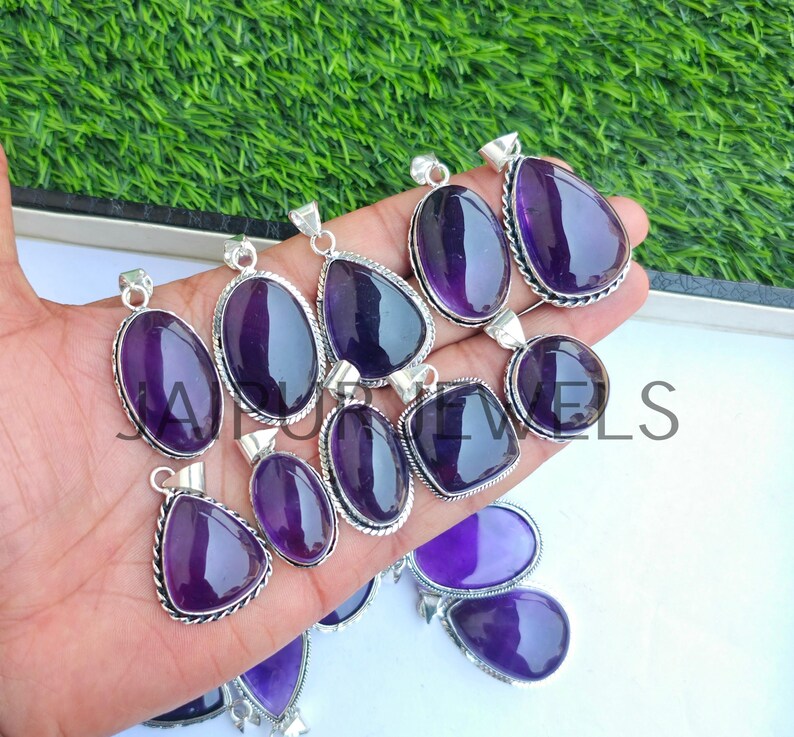 May include: A collection of amethyst pendants in various shapes, including oval, teardrop, and square. Each pendant features a polished, deep purple gemstone set in a silver-colored metal frame. The pendants are displayed against a green background.