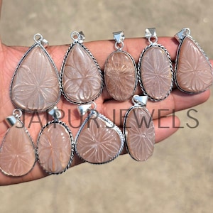 May include: A collection of light pink gemstone pendants in various shapes, including teardrop and oval, with intricate carved floral designs. Each pendant has a silver-colored bail and a decorative silver border. The pendants are displayed on a hand.