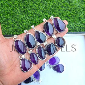 May include: A collection of amethyst pendants in various shapes, including oval, teardrop, and round. Each pendant features a polished, deep purple gemstone set in a silver-toned metal frame. The pendants are displayed against a green background.