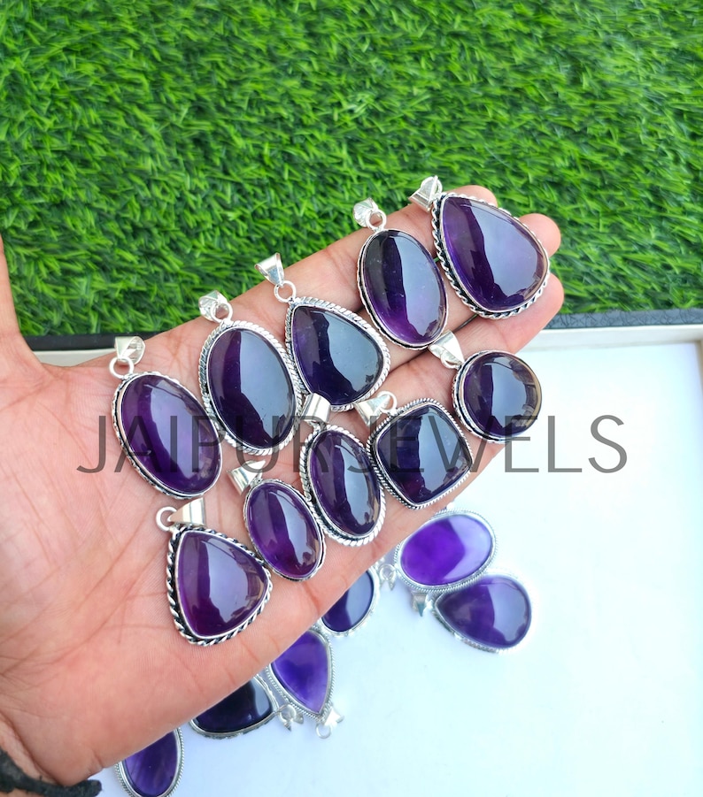 May include: A collection of amethyst pendants in various shapes, including oval, teardrop, and round. Each pendant features a polished, deep purple gemstone set in a silver-toned metal frame. The pendants are displayed against a green background.