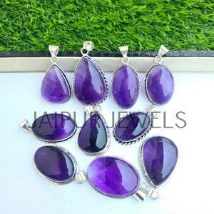May include: A collection of amethyst pendants in various shapes, including teardrop and oval. Each pendant features a polished, deep purple gemstone set in a silver-colored metal frame. The text "JAIPUR JEWELS" is visible.