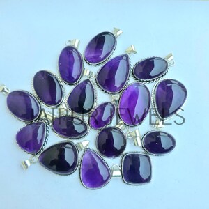 May include: A collection of amethyst pendants in various shapes, including oval, teardrop, and square. Each pendant features a polished, deep purple gemstone set in a silver-toned metal frame. The pendants are displayed on a light background.