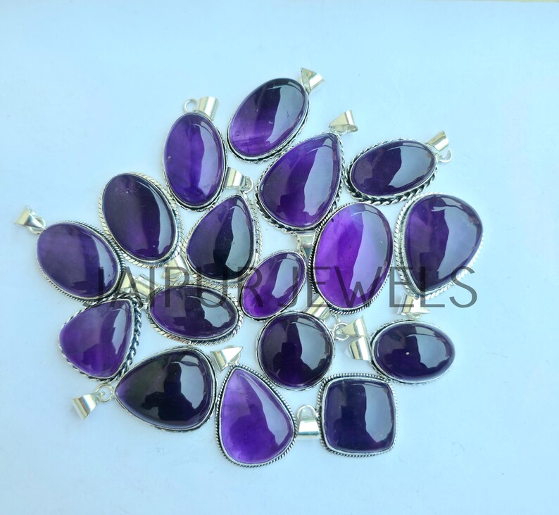 May include: A collection of amethyst pendants in various shapes, including oval, teardrop, and square. Each pendant features a polished, deep purple gemstone set in a silver-toned metal frame. The pendants are displayed on a light background.