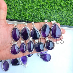 May include: A collection of amethyst pendants in various shapes, including oval, teardrop, and square. Each pendant features a polished, deep purple gemstone set in a silver-toned metal frame. The pendants are displayed against a green and white background.