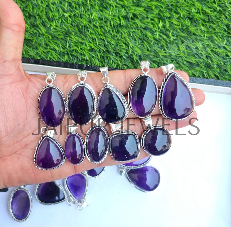 May include: A collection of amethyst pendants in various shapes, including oval, teardrop, and square. Each pendant features a polished, deep purple gemstone set in a silver-toned metal frame. The pendants are displayed against a green and white background.