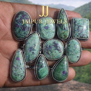 Peut inclure: Une collection de bagues en pierres précieuses de différentes formes, dont ovales, en goutte et carrées. Les bagues présentent des pierres vertes et violettes serties dans du métal argenté. Le texte "JAIPUR JEWELS" est visible.