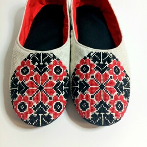 May include: A pair of white linen slippers with red and black embroidered floral designs. The slippers have a black lining and a red trim around the top.
