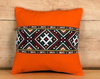 Puede incluir: Cojín naranja con una banda bordada en negro, blanco, verde y rojo con un patrón geométrico.