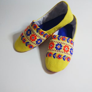 May include: A pair of yellow embroidered slippers with blue and red floral patterns. The slippers have a blue lining and are made of a soft, comfortable fabric.