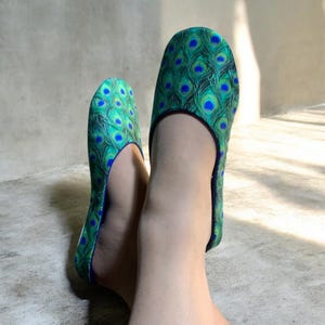 May include: A pair of slip-on shoes featuring a vibrant peacock feather pattern in shades of emerald green, teal, and royal blue. The footwear has a rounded toe design with a thin blue trim along the opening, providing a snug fit around the foot.