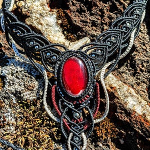 May include: A black macrame necklace with a red gemstone pendant. The necklace is made with black cord and silver beads. The pendant is a large, oval-shaped gemstone with a deep red color.