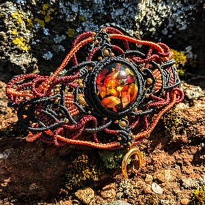 May include: A black, red, and orange macrame bracelet with a gold crescent moon charm and a round resin cabochon featuring pressed flowers.