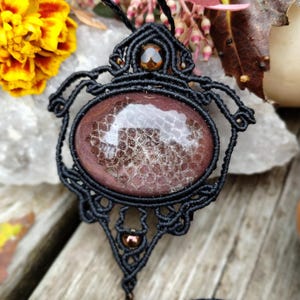 May include: A black macrame pendant featuring a large, round, brown and white agate stone. The pendant is accented with small, brown beads.