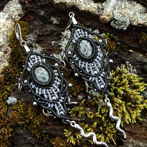 Peut inclure: Une paire de boucles d'oreilles macramé faites à la main. Chaque boucle d'oreille présente une pierre ovale centrale, entourée de macramé noir et gris complexe. Des perles argentées et un charme de serpent argenté complètent le design. Les boucles d'oreilles sont présentées sur une surface texturée et moussue.