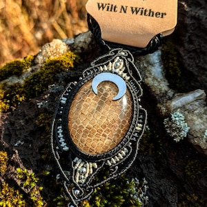 以下が含まれることがあります： 三日月形のチャームと茶色のテクスチャのある石のペンダントが付いた黒いマクラメネックレス。ネックレスに付けられたタグには「Wilt N Wither」と書かれています。