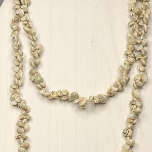 May include: A long, layered necklace made of small, beige seashells. The necklace is displayed on a white wooden jewelry stand. The shells have a natural, textured appearance, and the necklace is draped in a flowing, elegant style.
