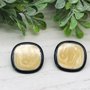 May include: Two square stud earrings with a marbled beige and gold design, set in a black frame. The earrings are displayed on a white surface with green foliage in the background.
