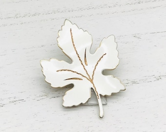 May include: White enamel leaf brooch with gold detailing. The leaf has a simple design with five points and a stem.