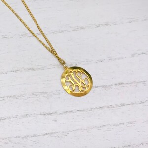 May include: A gold-colored necklace with a delicate chain and a circular pendant. The pendant features an intricate, openwork design of intertwined letters. The necklace is displayed on a white, wood-grain surface, with floral accents in the background.