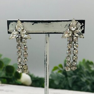 May include: A pair of vintage rhinestone earrings on a white display stand. The earrings feature a cluster of clear, teardrop-shaped stones at the top, with multiple strands of smaller, round rhinestones cascading downwards. The stand is white with a distressed finish.