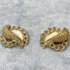 May include: A pair of gold-toned leaf-shaped clip-on earrings. Each earring features a textured leaf design with a detailed, openwork border. The earrings are set against a soft, gray background.