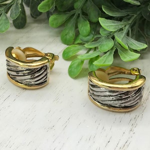 May include: A pair of clip-on earrings with a gold-tone frame and a silver-tone, ridged center. The earrings have a rounded, rectangular shape. The background includes green foliage and a white wooden surface.