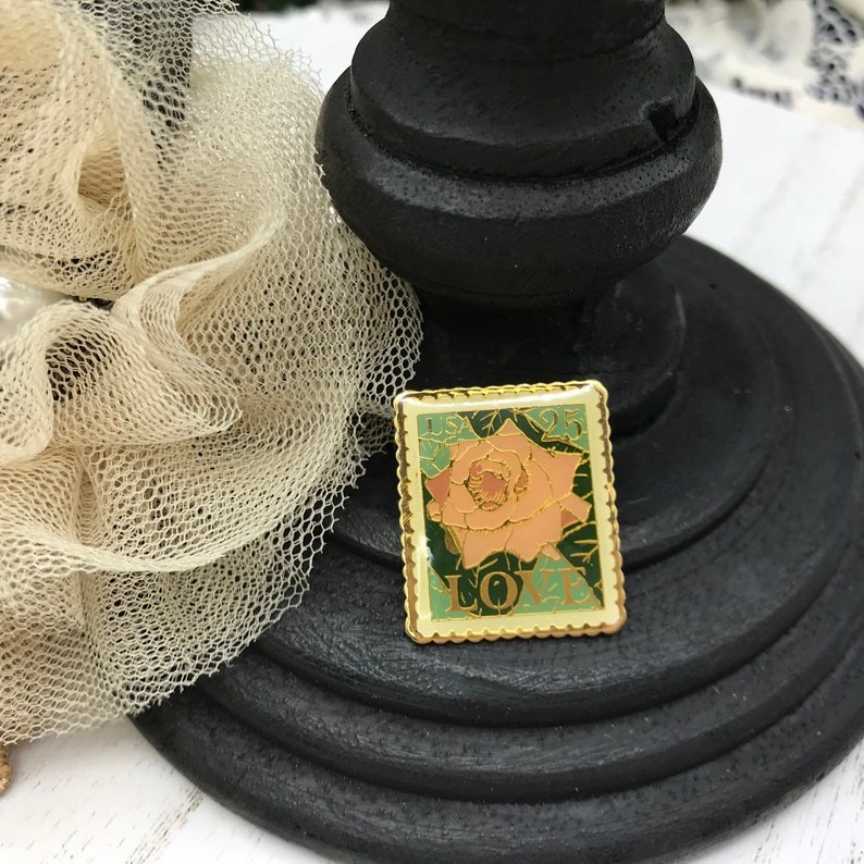 May include: A rectangular gold-edged pin with a rose design. The pin features a peach-colored rose with green leaves and the word "LOVE" in green. The pin also has the text "USA 25" in the upper corners. The pin is on a black stand.