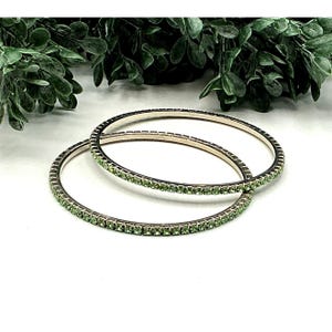 Puede incluir: Dos pulseras de plata con una fila continua de pequeñas piedras preciosas verde claro. Las pulseras están sobre un fondo blanco, con follaje verde en la esquina superior izquierda. Joyería sencilla y elegante.