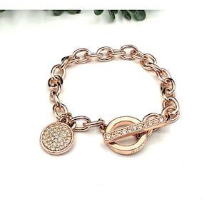 May include: Rose gold bracelet with a toggle clasp and a round charm. The charm and clasp are embellished with small, clear crystals. The bracelet has a chain link design and is displayed on a white surface.