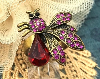 May include: A gold-tone ring with a red gemstone and a detailed design of a dragonfly with pink rhinestones.