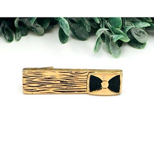 May include: Gold-toned tie bar with a black bow tie design. The tie bar has a textured surface.