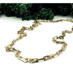 May include: A gold-colored chain necklace with rectangular and square links. The necklace is displayed on a white surface with green foliage in the background. The chain is made of metal and has a shiny finish.