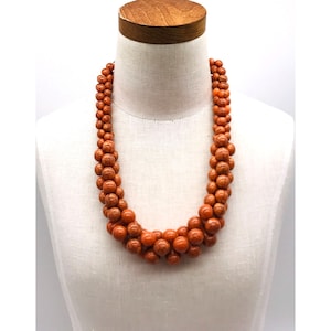 May include: A necklace composed of multiple strands of orange-red beads. The beads vary in size, creating a textured, layered effect. The necklace is displayed on a white mannequin, showcasing its design and color.